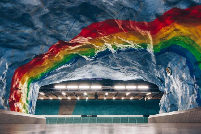 The hall of Stadion subway station. The rock ceiling is bright blue, with a rainbow painted across it.